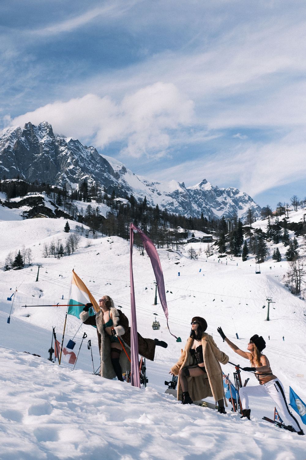 Three fashionable skiers pose with colorful ski poles on a snowy Alpine slope with dramatic mountain peaks behind them.