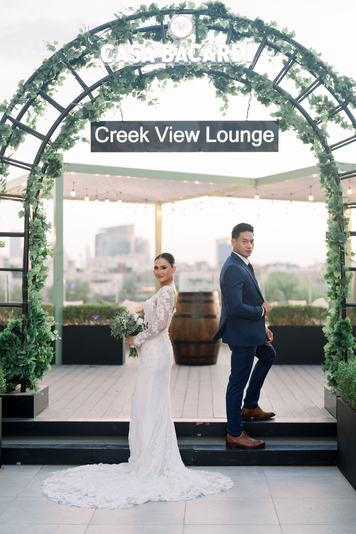Couple stands in elegant wedding attire under floral archway at Creek View Lounge with city skyline in background.