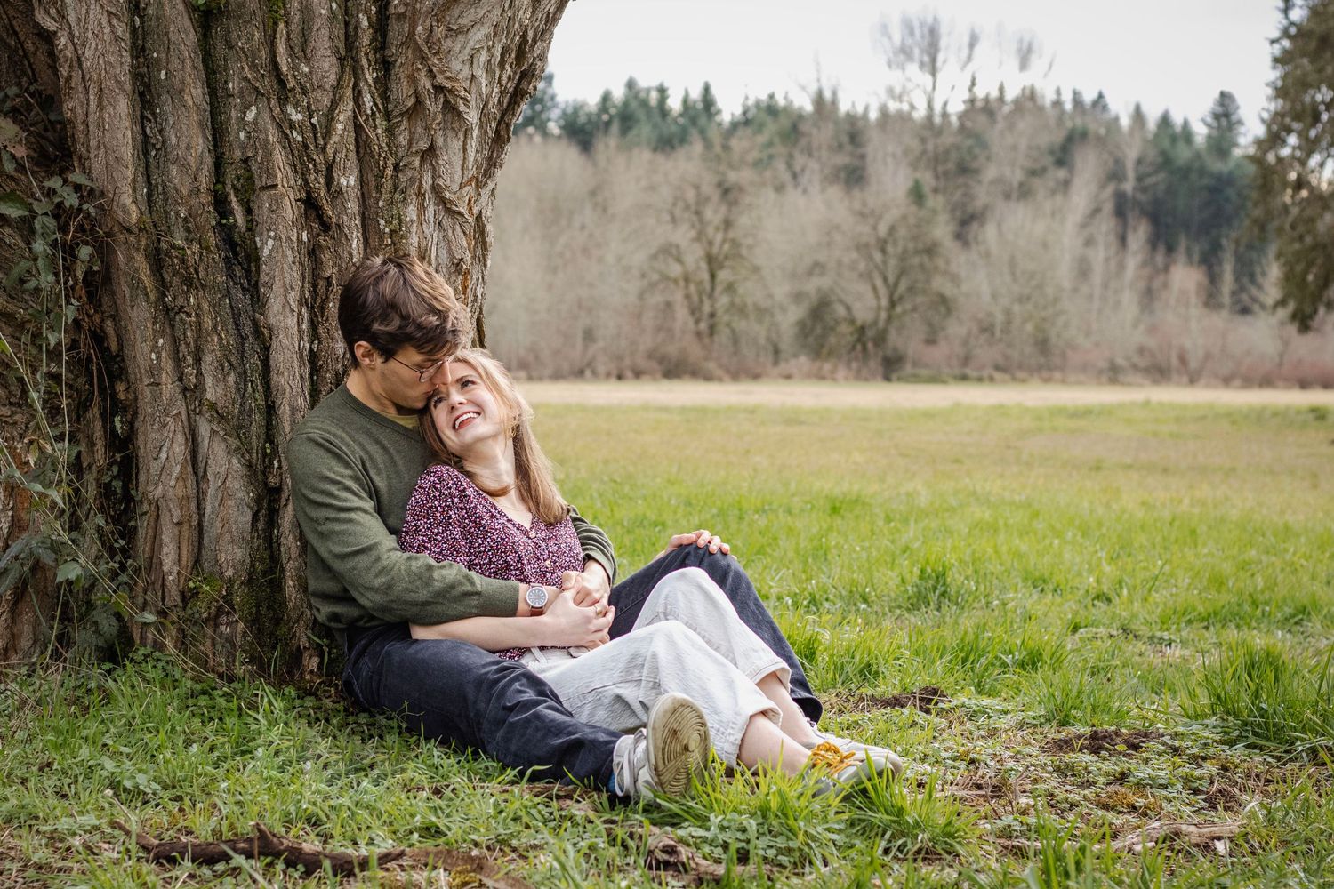 a young man and woman sit under a tree in a meadow. he hugs her while she looks back at him and smiles