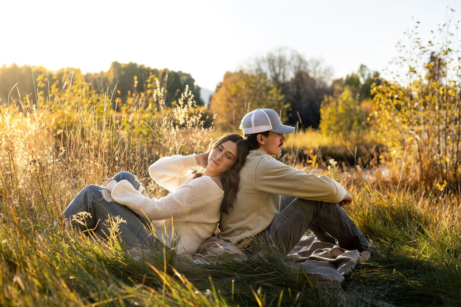 a young man and woman sit under a tree in a meadow. he hugs her while she looks back at him and smiles
