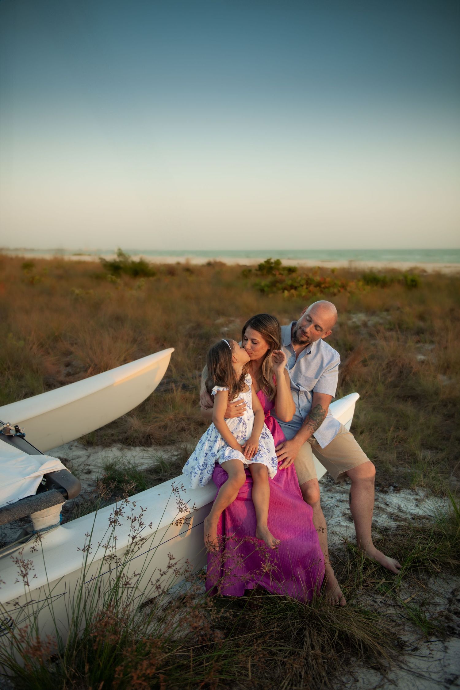 A family sits together on white surfboards at the beach during sunset with tall grass and pink floral details in view.