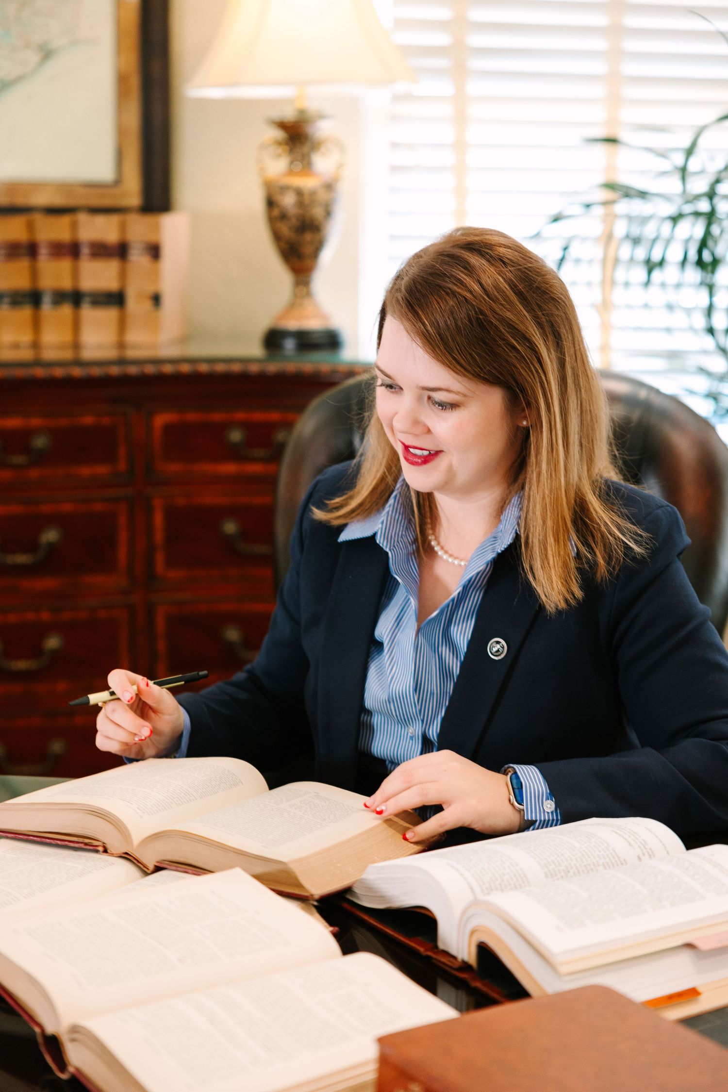 Professional reading reference books at a wooden desk with a table lamp and window blinds.