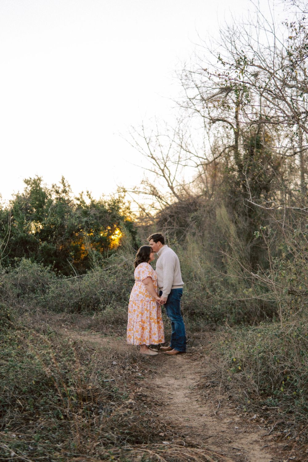 A couple shares an intimate moment on a natural dirt path surrounded by trees and brush at sunset.
