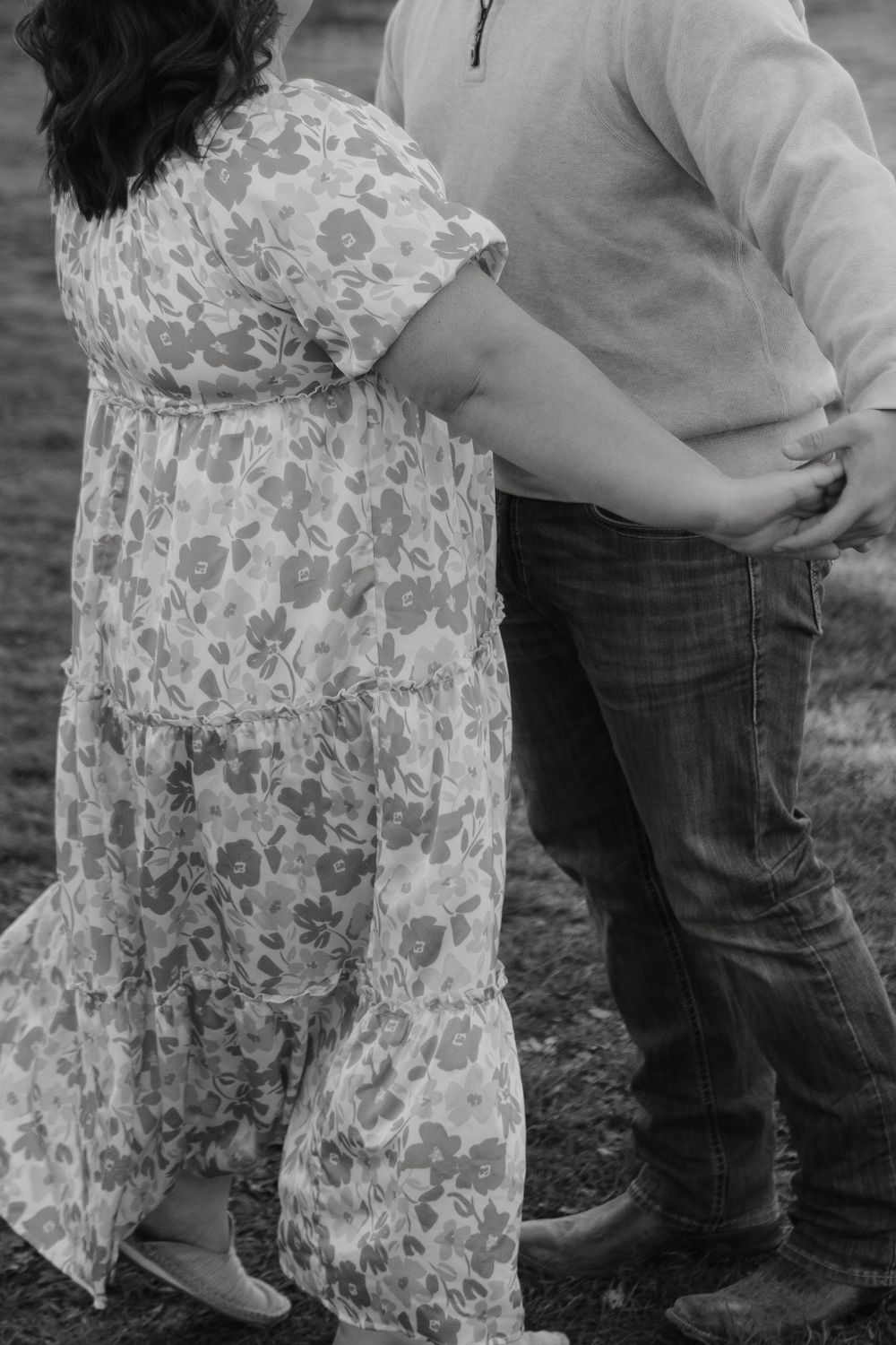 Black and white photo showing a couple standing close together, with a floral dress and jeans visible against a grassy backdrop.