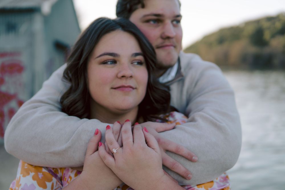 A couple embraces outdoors near the water on a cloudy day, wearing casual clothing.