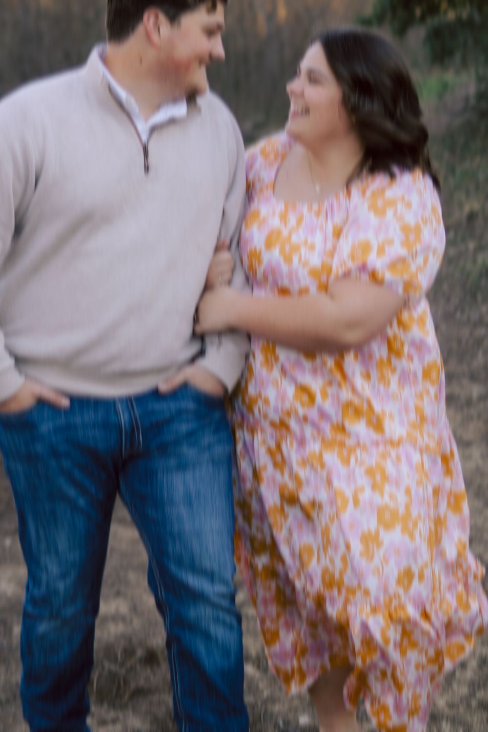 A couple shares a candid laughing moment together outdoors, with her wearing a flowing floral dress and him in jeans and sweater.