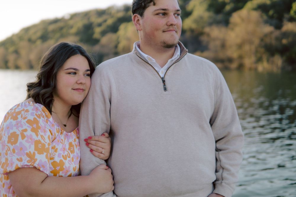 A couple stands close together by a lake at sunset wearing casual attire with autumn foliage in the background.