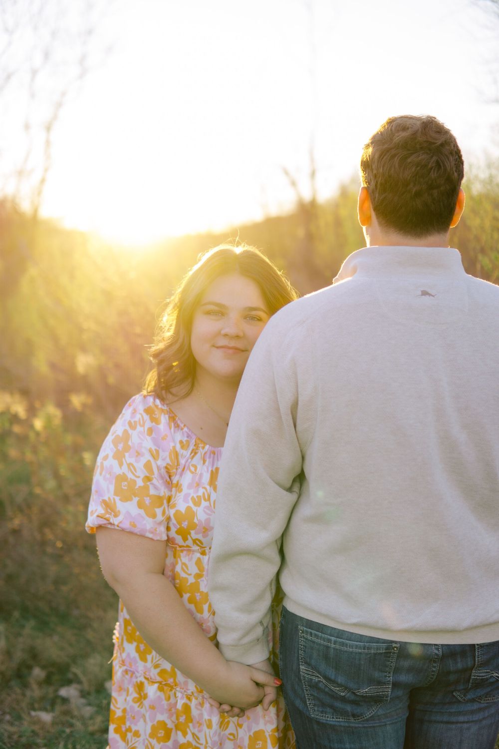 A couple stands together in a field at golden hour, with sunlight creating a warm backlit glow through the trees.