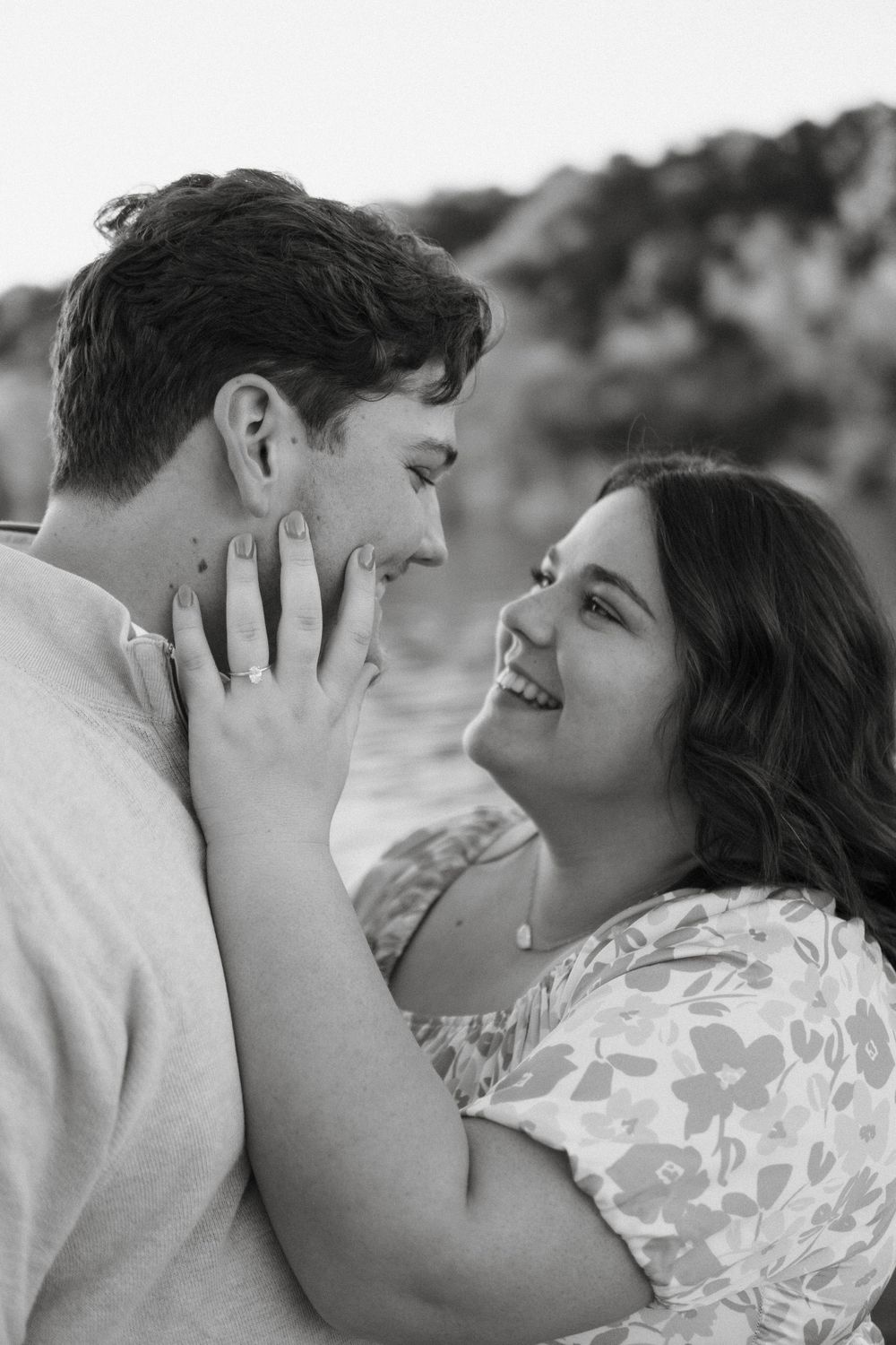 A romantic black and white portrait of a couple sharing an intimate moment outdoors while smiling at each other.