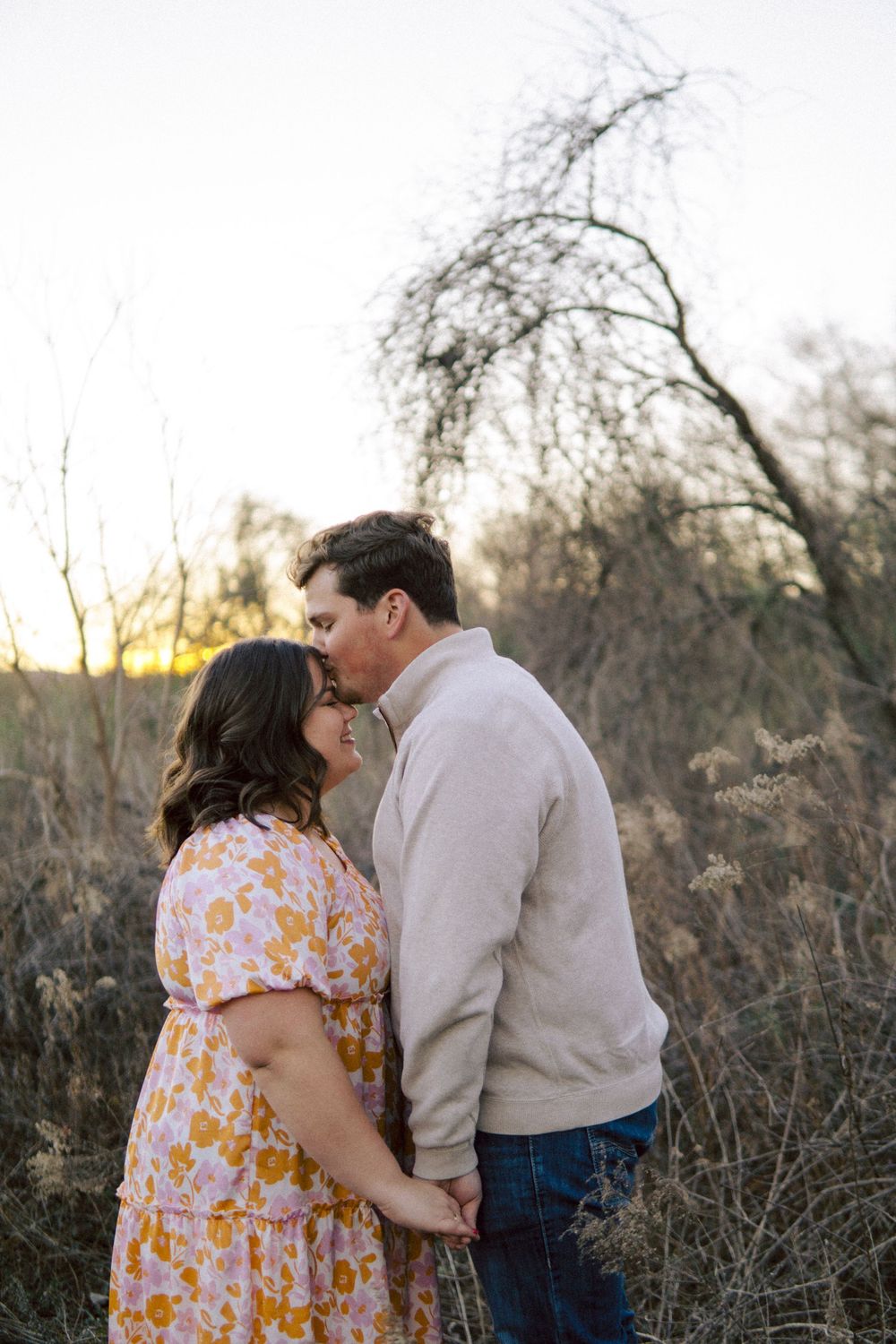 Romantic couple embraces in a floral dress and beige sweater against a naturalistic autumn backdrop at sunset.