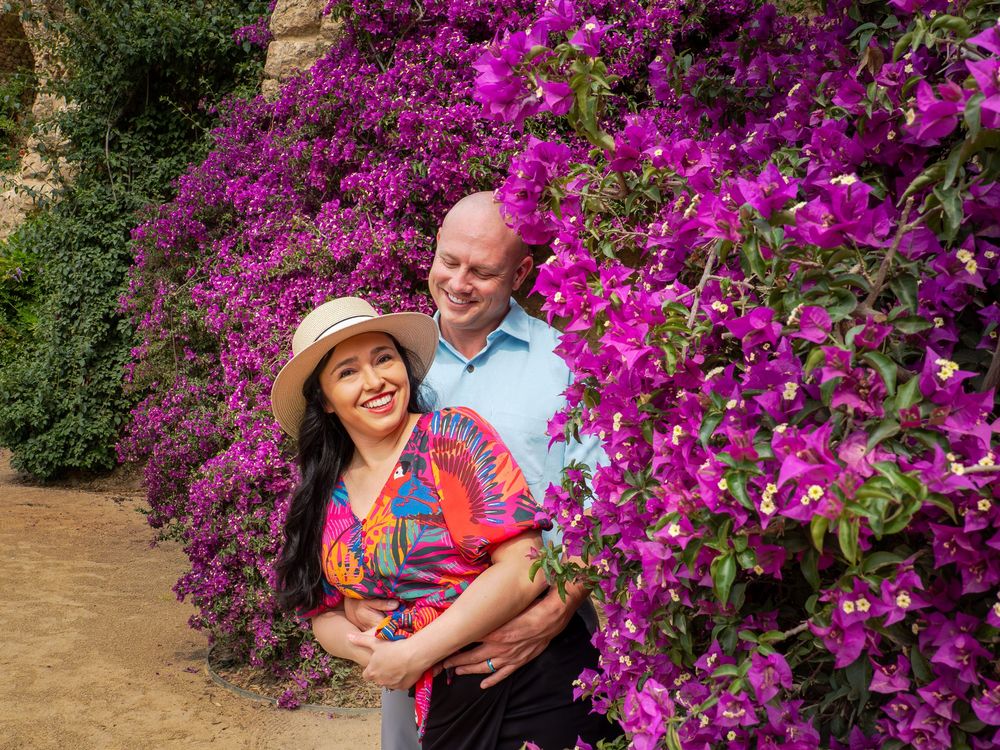 Couple smiling among vibrant purple bougainvillea flowers, woman in colorful top and hat, man in blue shirt.