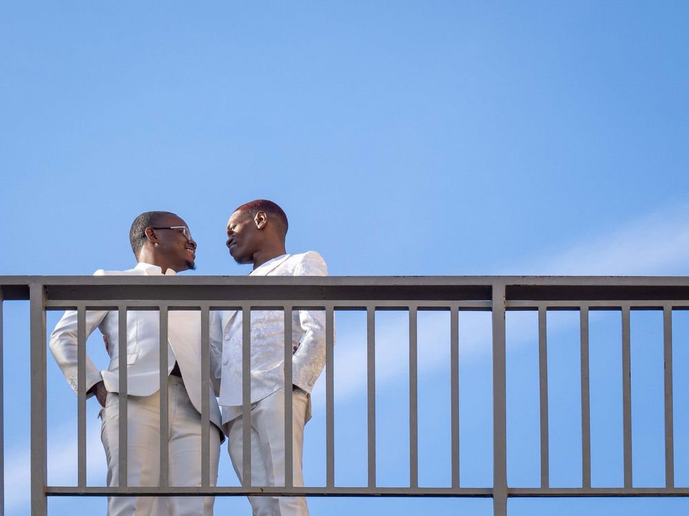 Two men in white suits stand talking on a bridge against a clear blue sky.