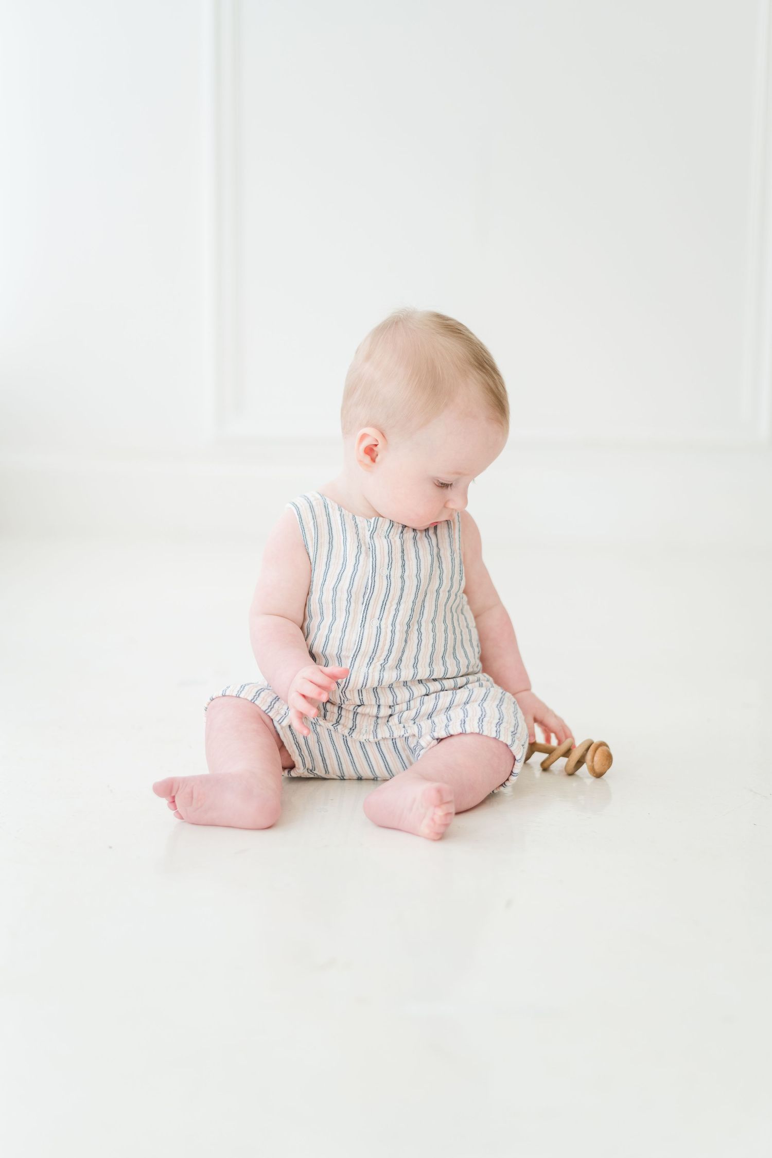 An adorable baby in a striped romper sits on a white floor playing with a small wooden toy.