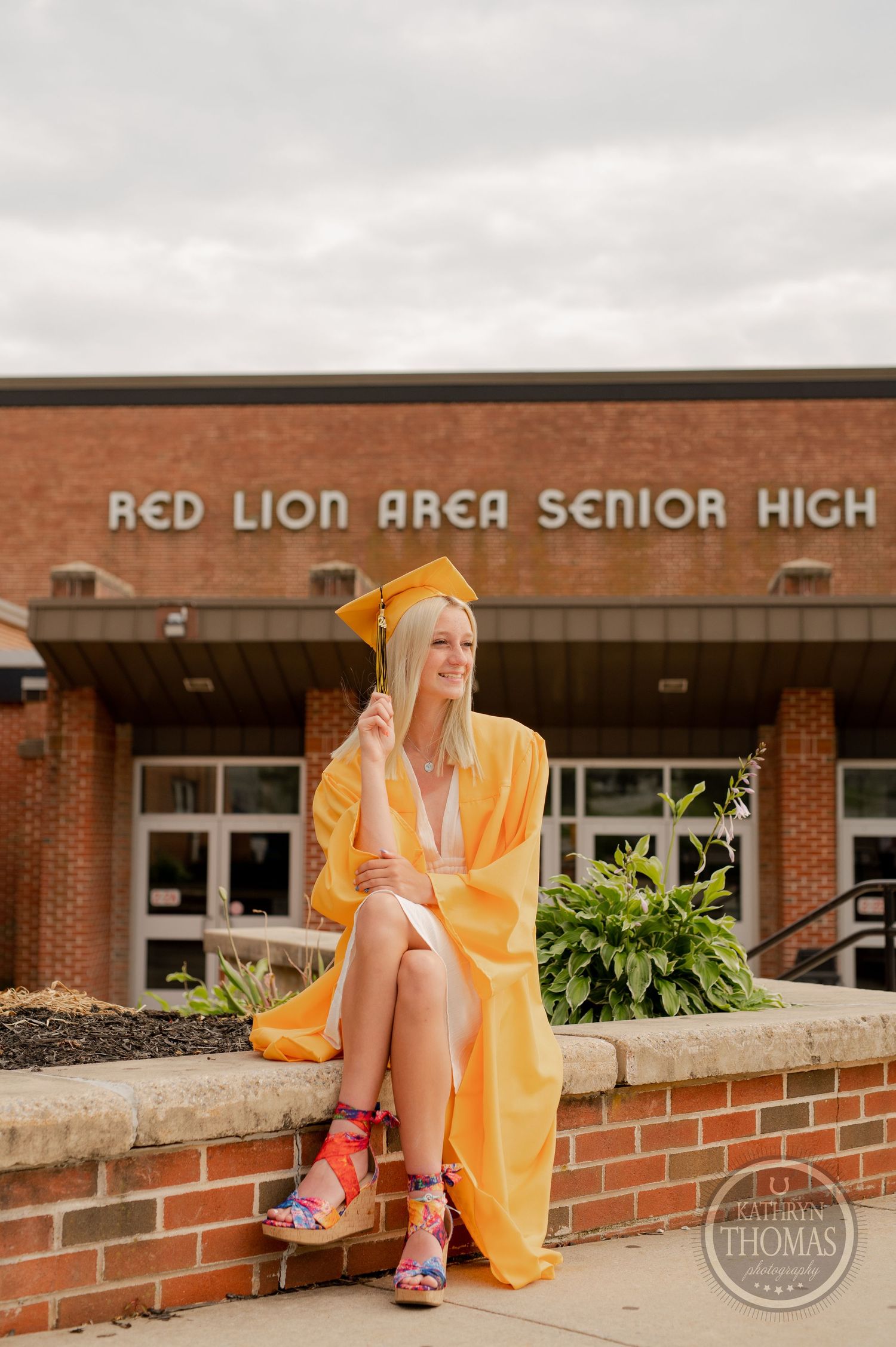 Graduate in yellow graduation attire poses on brick wall wearing colorful sneakers.