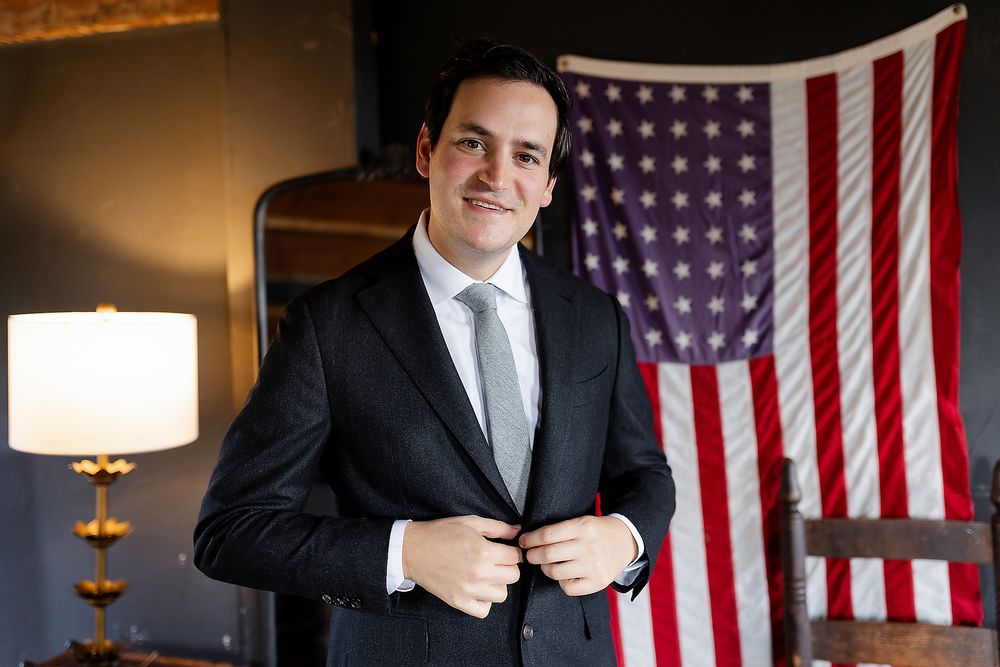A formal suit against an American flag backdrop in an interior setting.