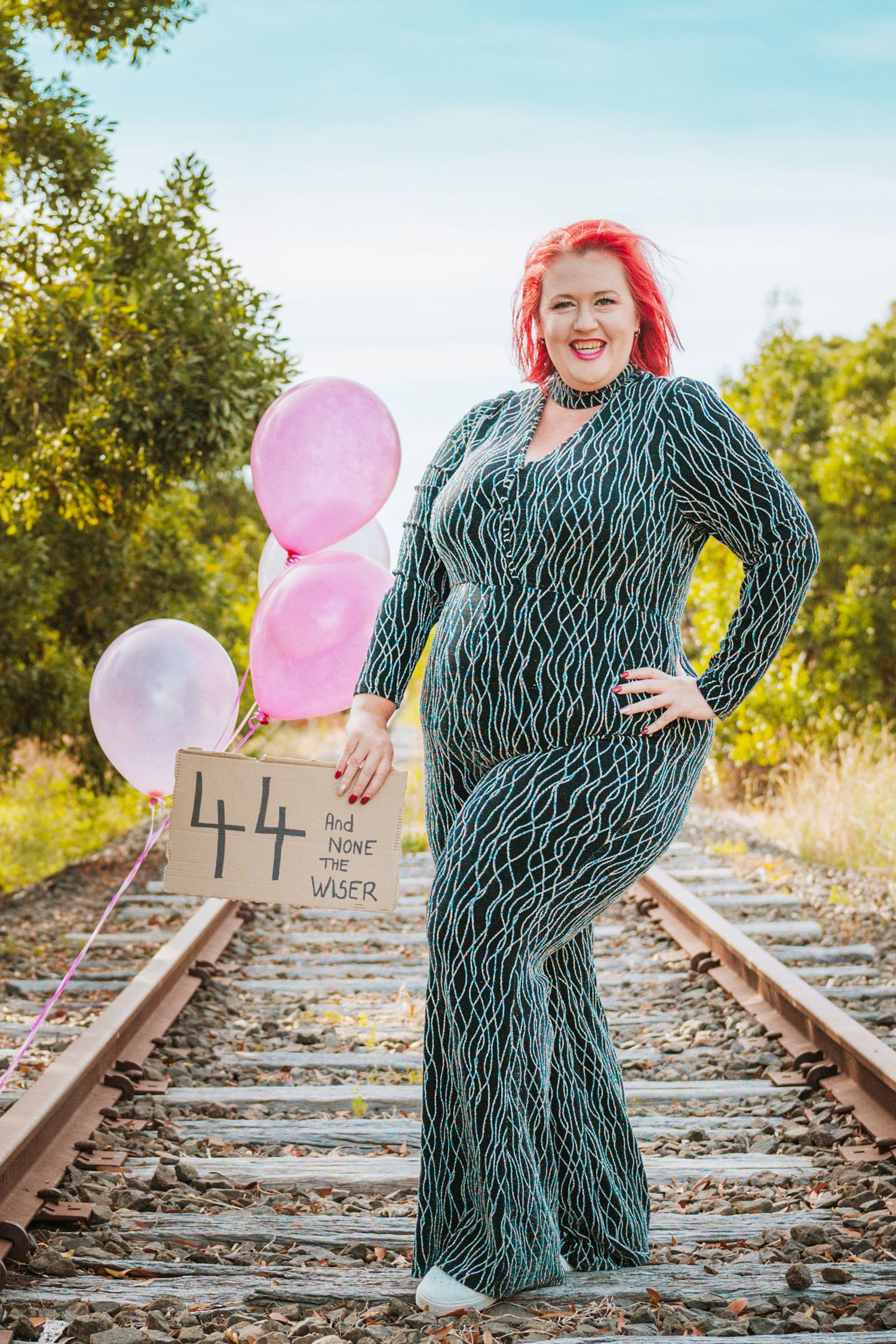 A person in a patterned dress poses on train tracks at sunset with pink balloons and train signals in the background.