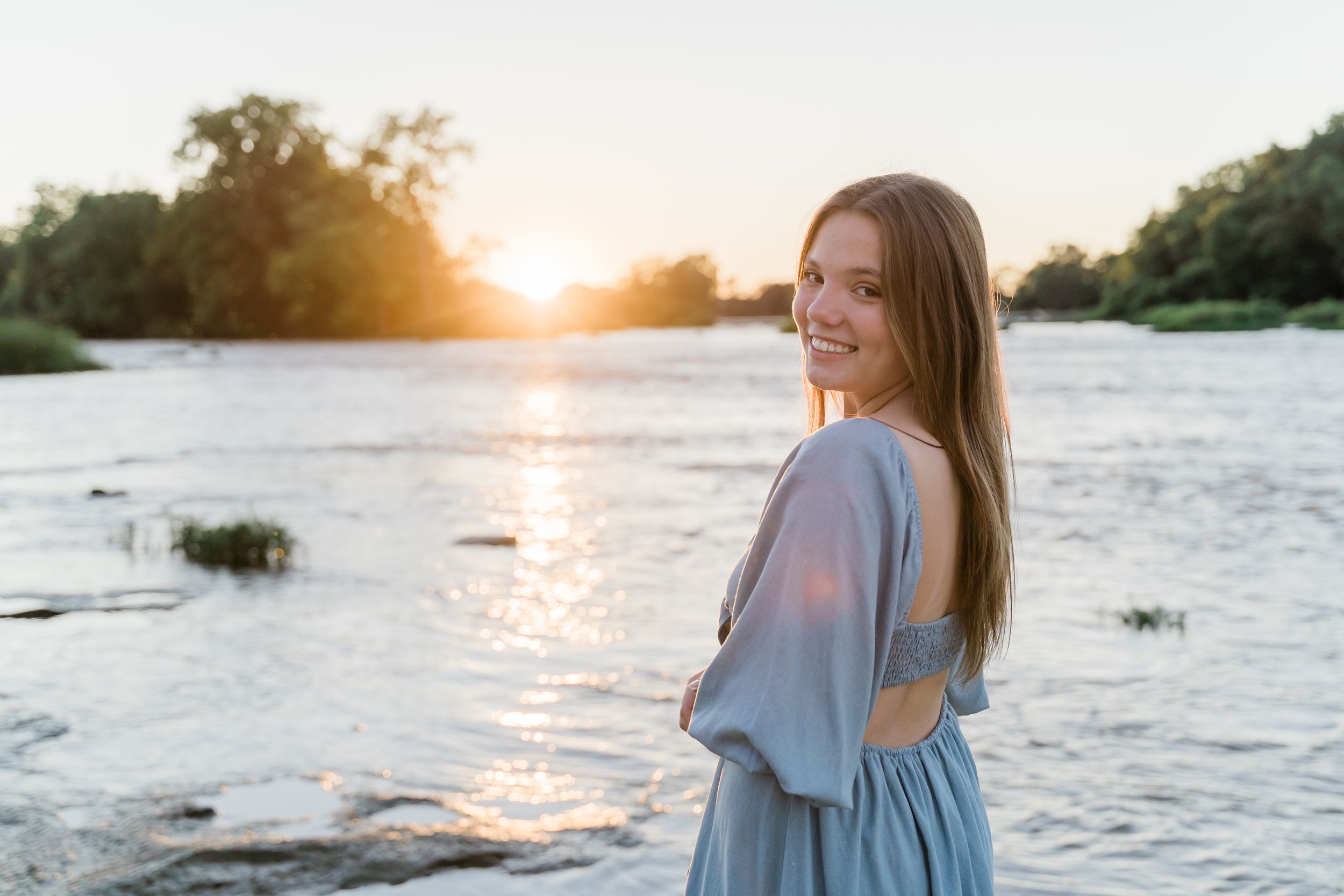 Woman in a light blue dress stands at the water's edge during sunset creating a serene lakeside scene.