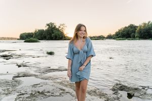 A person in a blue dress walks along a rocky riverbank during sunset with calm water and trees in background.