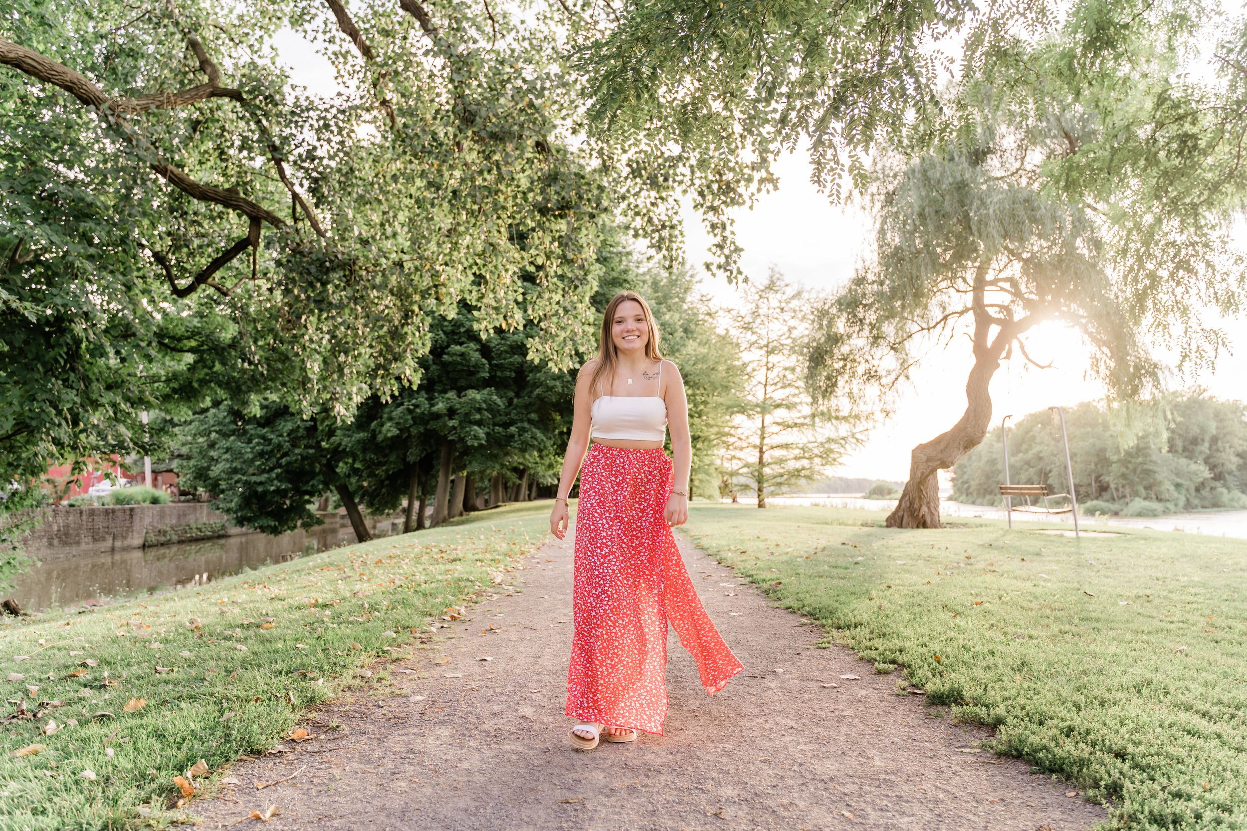 A person in a coral maxi dress walks along a tree-lined path during golden hour sunset in a park setting.