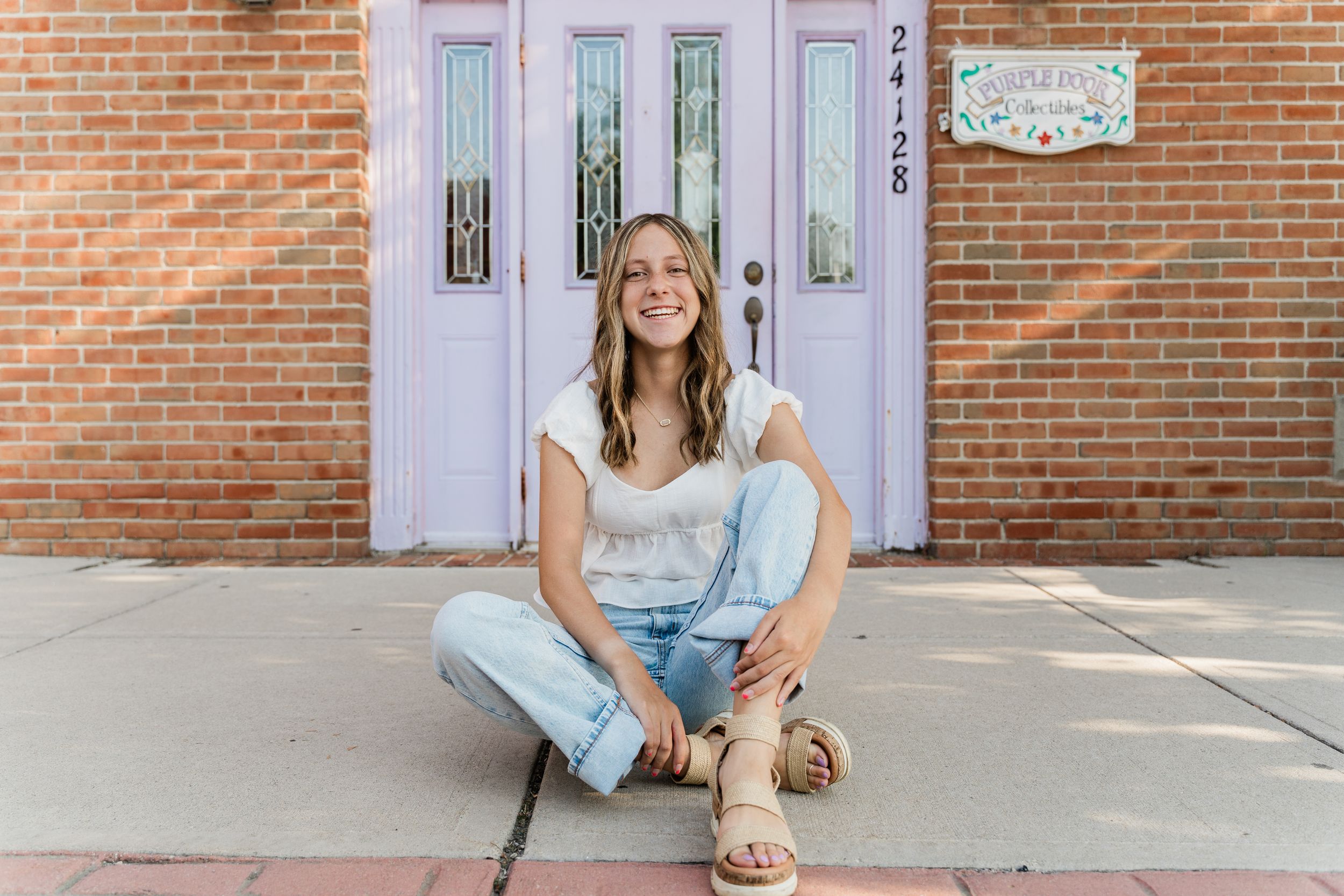 A woman with blonde hair in a blue sweater sits on a white porch step outside a brick building.
