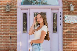 Woman in blue overalls and white top poses in front of a blush pink door with brick wall background.