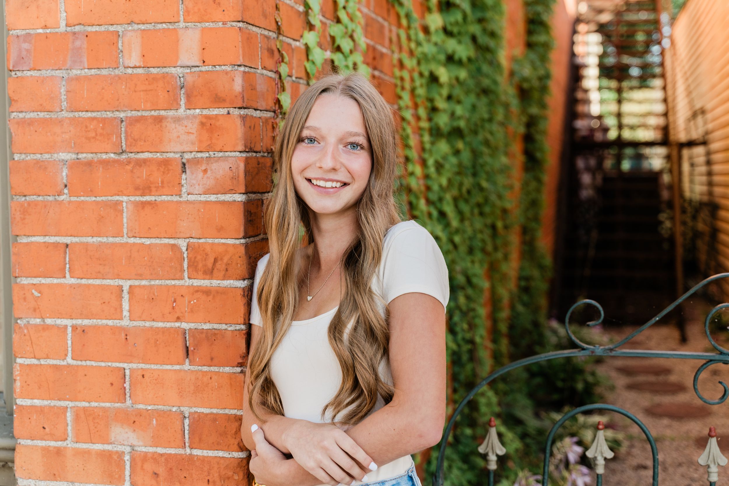 A person in a white t-shirt smiles while standing against an orange brick wall with ivy growing in the background.