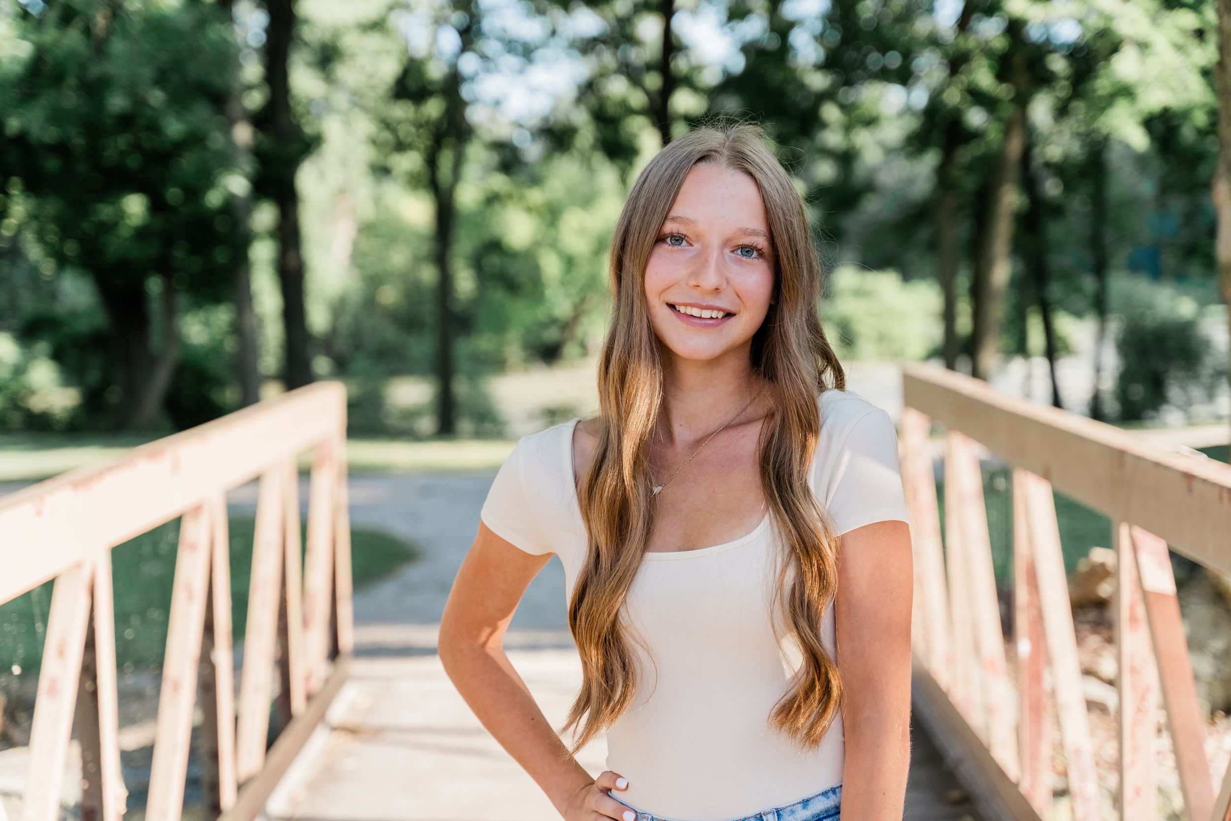 Young woman in white top and denim shorts standing on wooden bridge in forest setting during sunny day.
