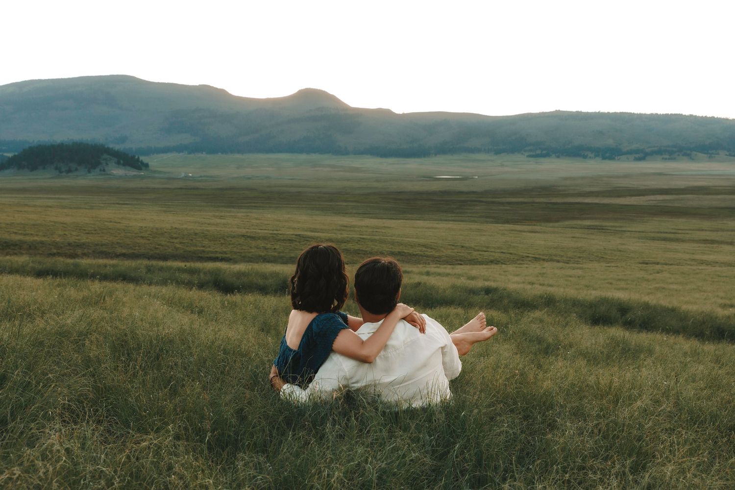 A couple sits together in tall grass gazing at a serene mountain valley landscape at dusk.