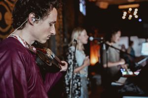 A musician playing violin during a live performance with band members in background.