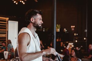 Performer in white tank top speaking at dimly lit event venue.