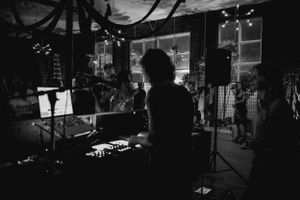 DJ silhouette performs at a dimly lit venue with electronic equipment and stage lighting.