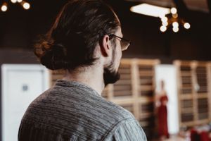 Person with glasses and man bun hairstyle shown from behind in an indoor setting with ambient lighting.