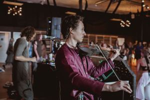 A violinist performing in a burgundy shirt with people dancing in the background.