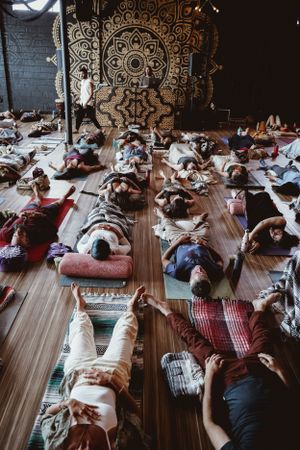 Large group practicing meditation on yoga mats in a studio with ornate mandala wall art.