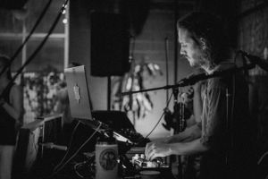 Artist performs on electronic music equipment in moody black and white lighting.