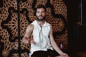 Wellness instructor in white clothing seated against ornate mandala wall design.