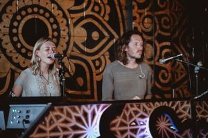 Musicians sing into microphones against a decorative patterned backdrop with illuminated designs.
