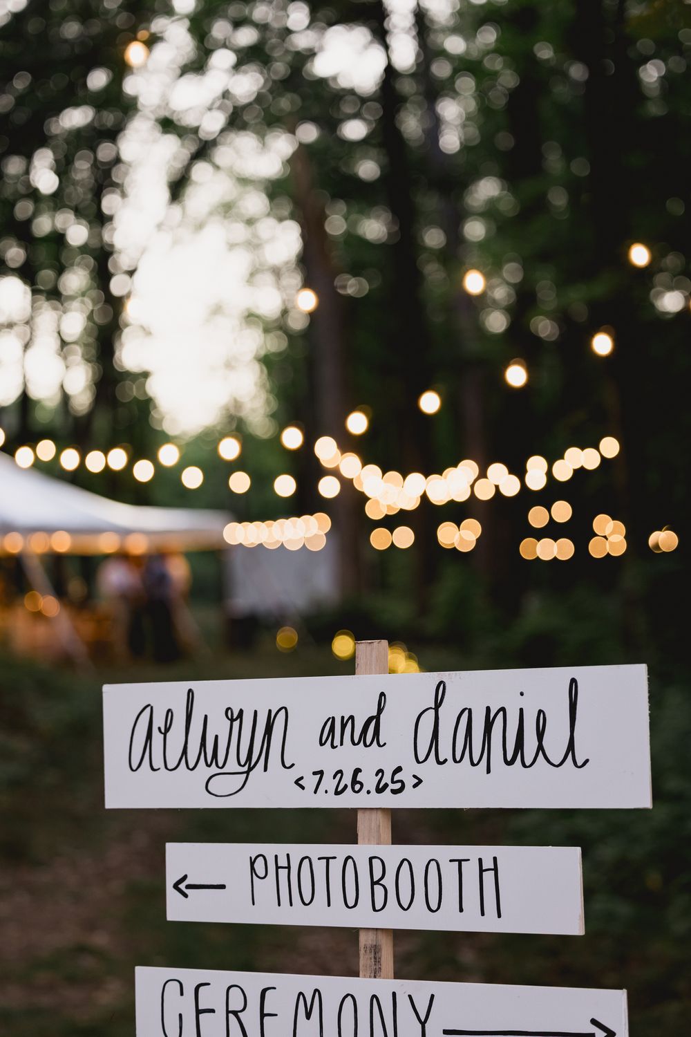 A wedding directional sign with string lights and bokeh effect in a forest setting at dusk.