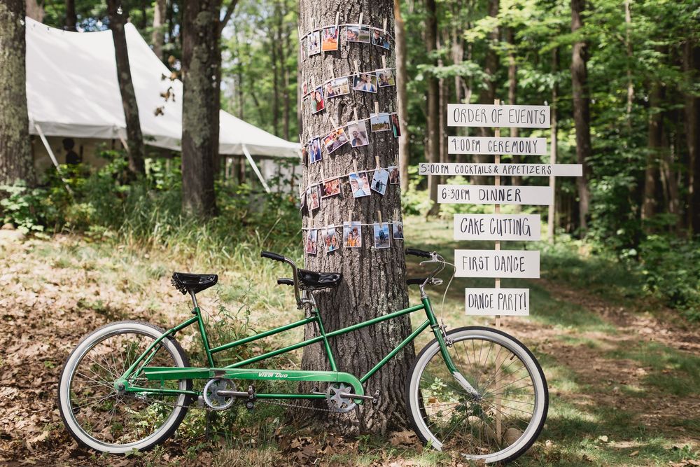 A vintage green tandem bicycle parked against a tree decorated with directional wooden signs in a forest setting.