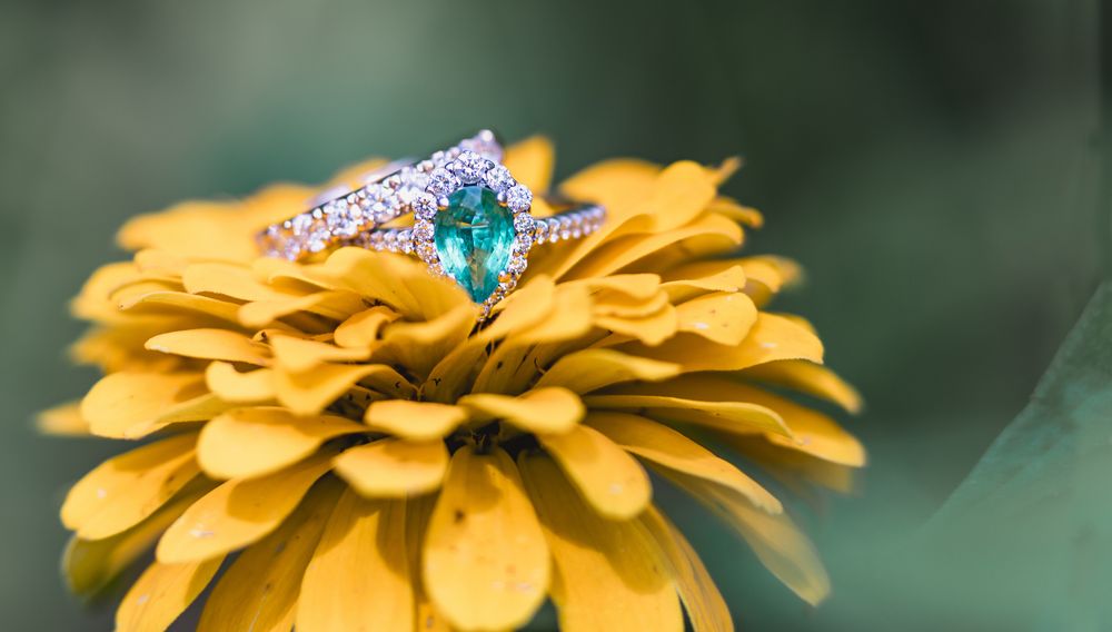 Diamond and emerald engagement ring resting on a bright yellow zinnia flower.