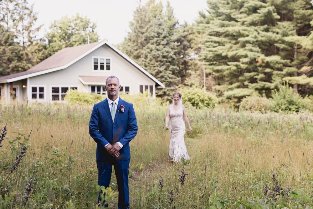 A couple stands in a field in front of a white house with a navy blue suit and white dress.