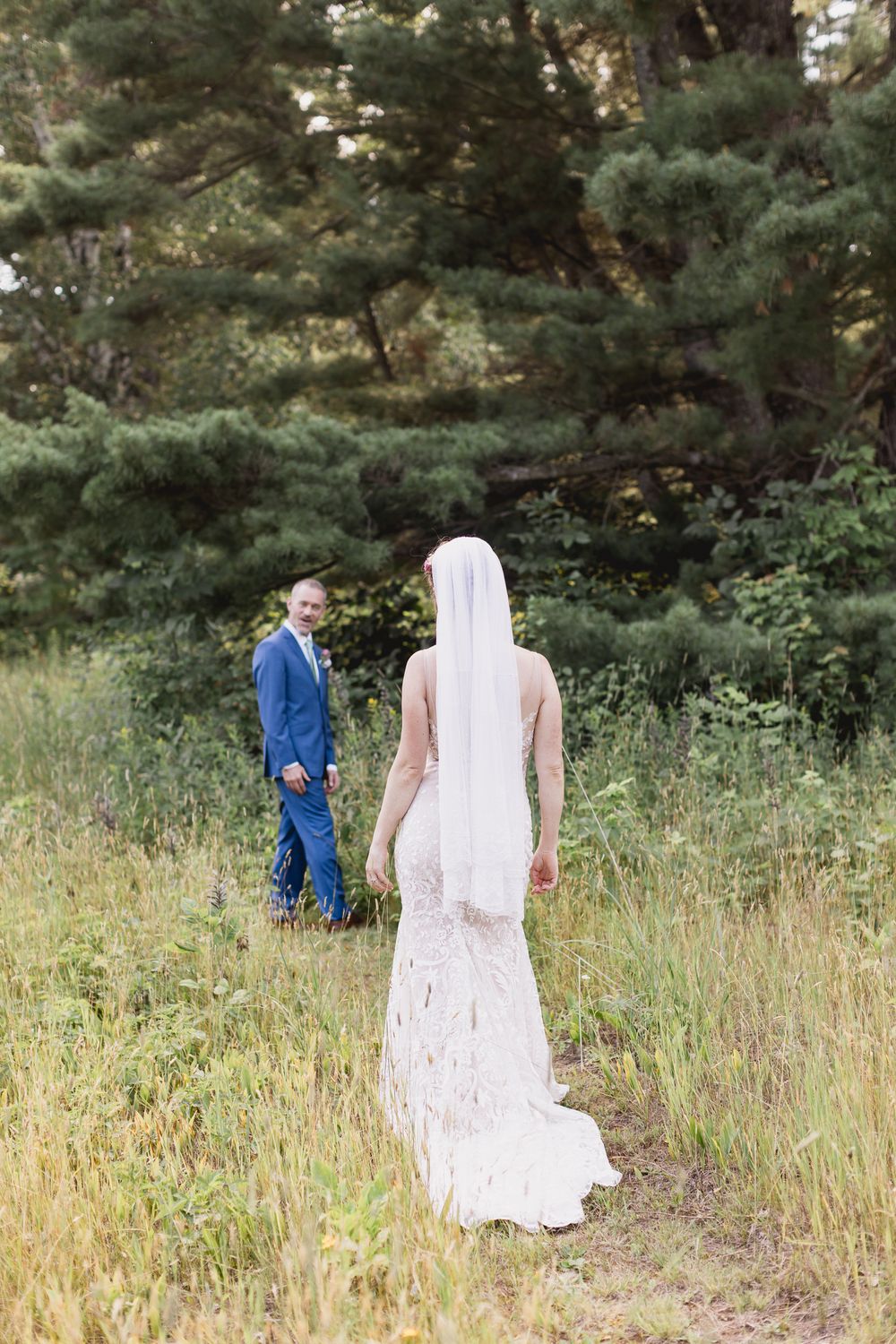 A couple shares a tender moment in a field with evergreen trees, the bride wearing a long lace gown and the groom in blue.