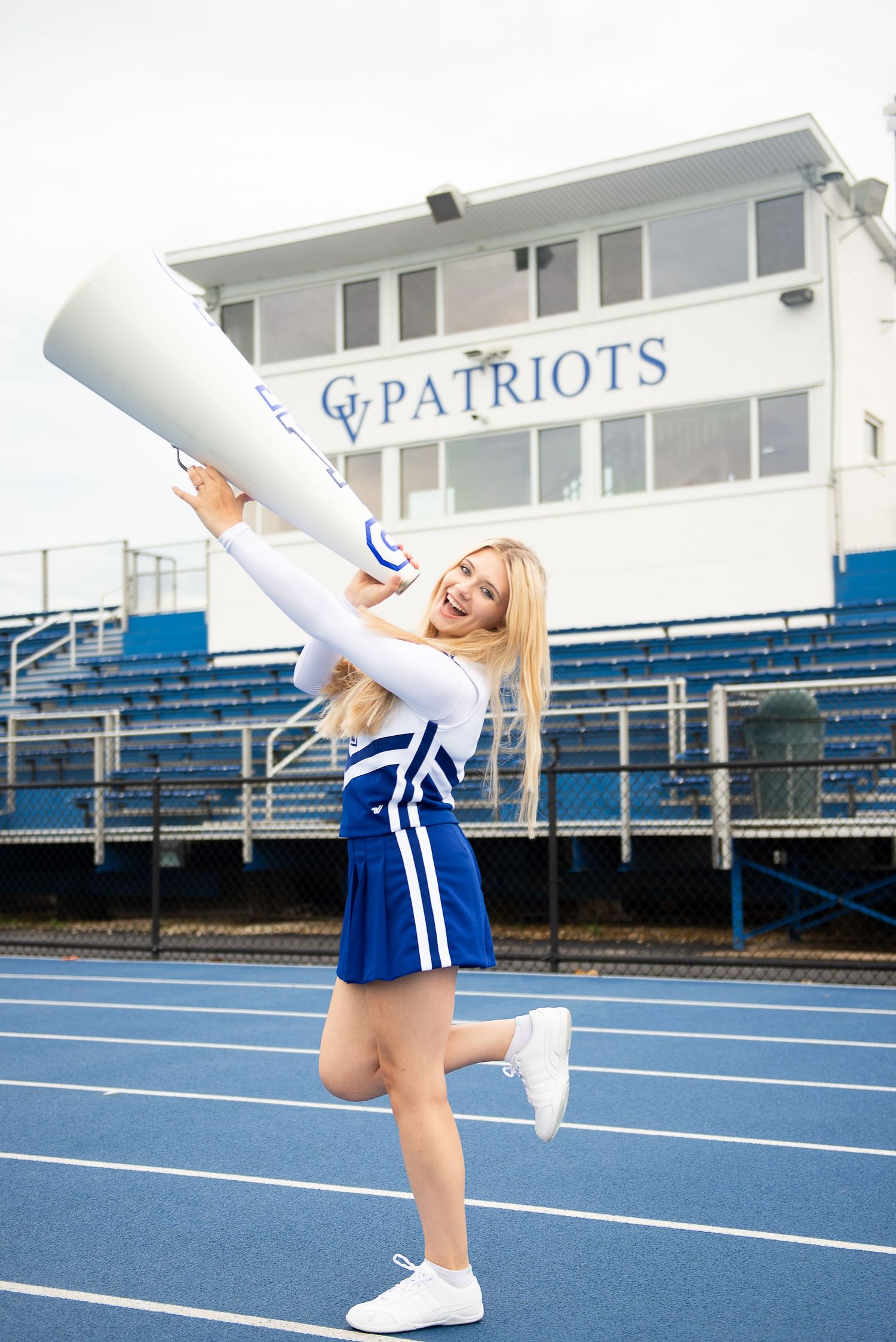 Cheerleader in blue and white uniform performs dynamic splits jumps on an outdoor athletic track at stadium.