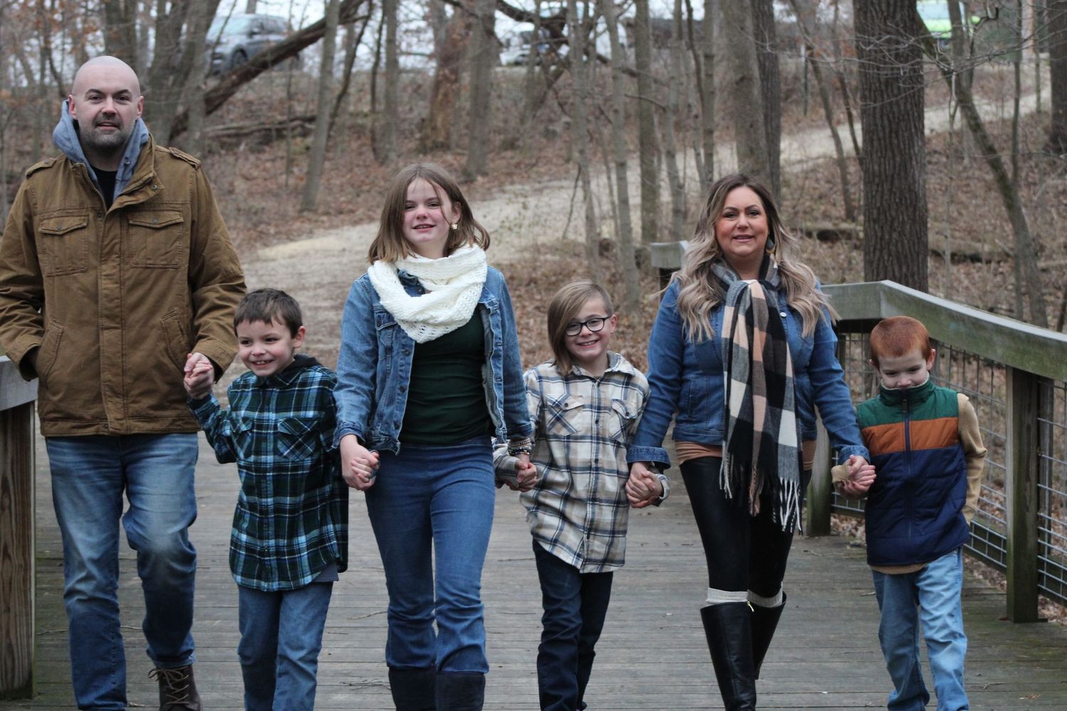 A family walk together along a wooden boardwalk path through a wooded winter landscape.