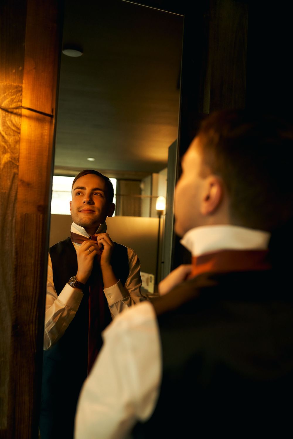 Someone adjusts their bowtie while looking in a mirror with warm atmospheric lighting.