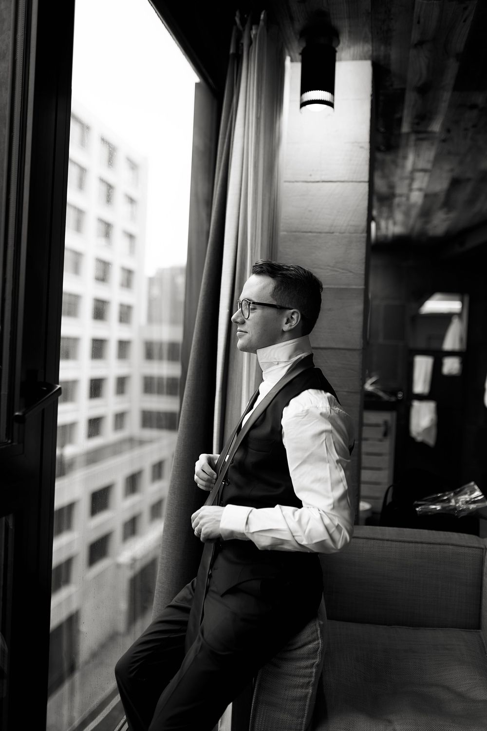 A couple in formal attire stands by a window in an intimate black and white portrait.