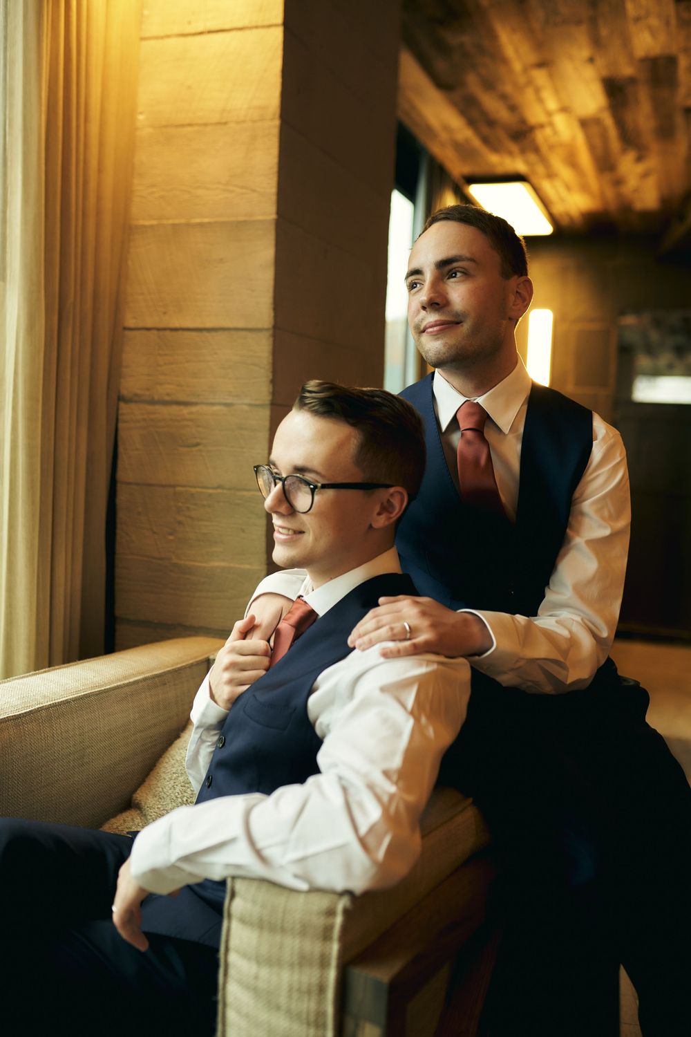 Two people in formal attire sit together by a window in warm lighting.