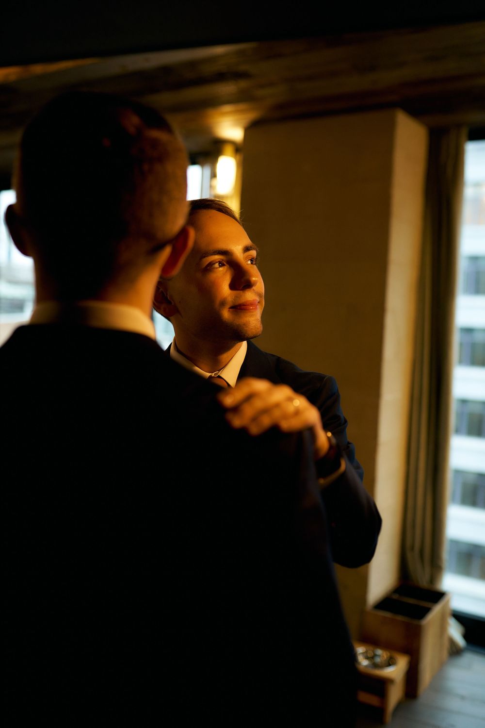 Two people in formal suits dancing together in warm lighting near windows.