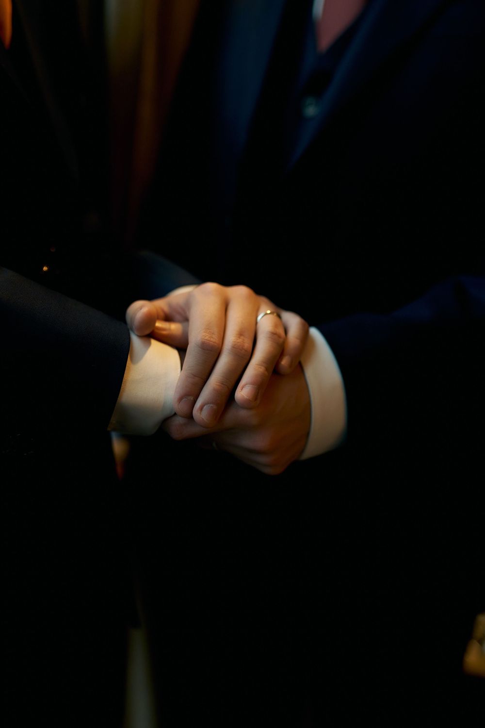Close-up of clasped hands showing wedding rings in dark formal attire.
