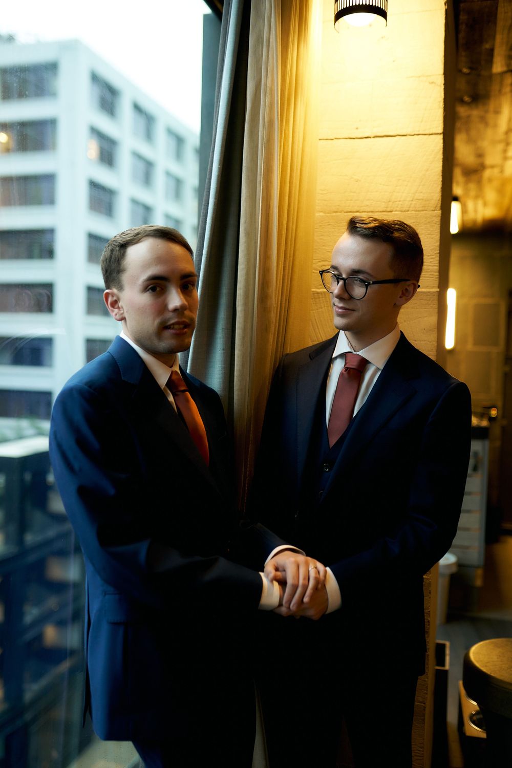 Two people in navy suits and red ties holding hands by a window in an urban setting.